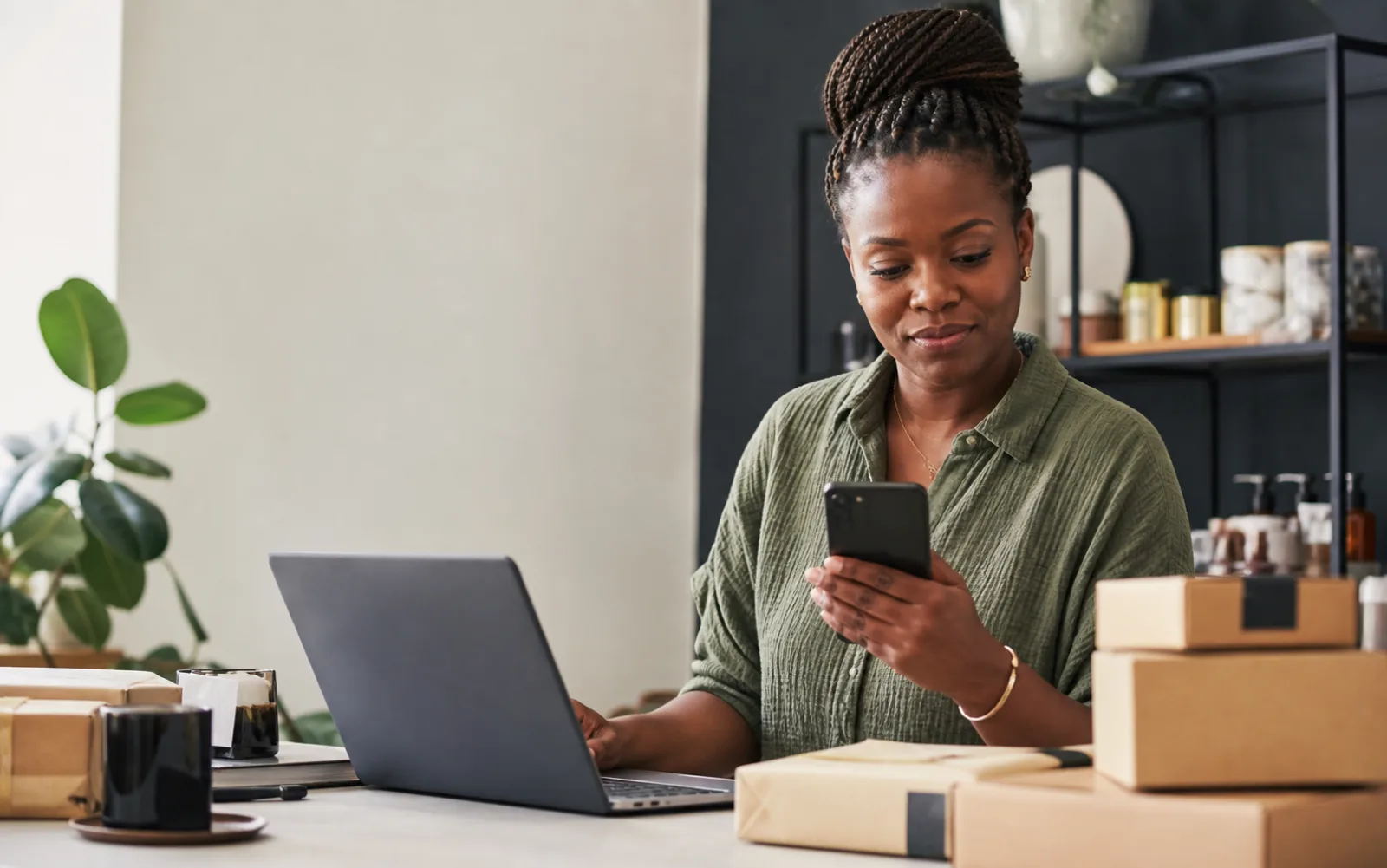 A Black ecommerce founder checking customer updates from her phone beside a laptop and parcels